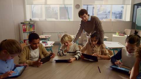 Enam siswa muda dengan tablet duduk di meja ruang kelas bersama, dan guru melihat tugas yang sedang dikerjakan dari belakang pundak siswa